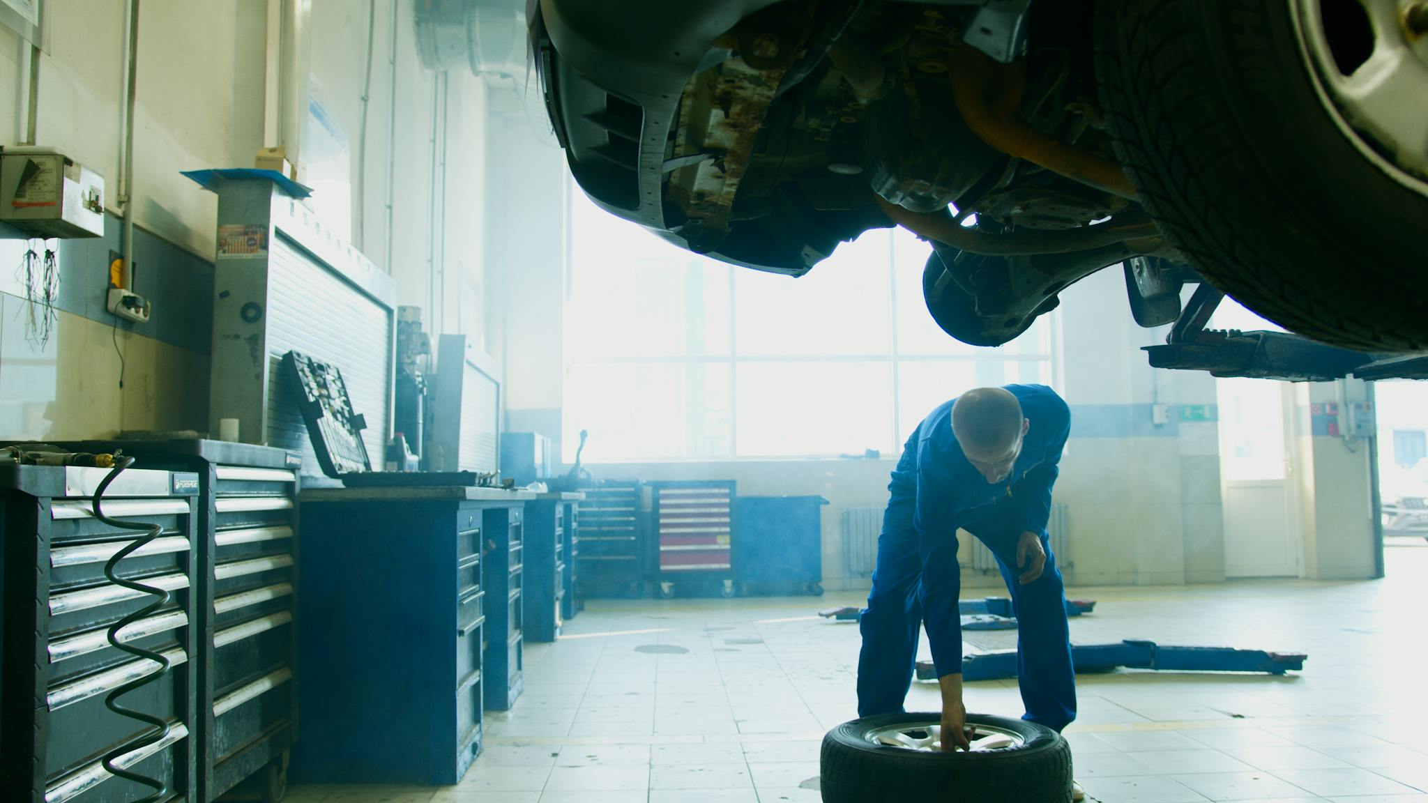 Mechanic in blue coveralls checking a tire under a vehicle in an indoor garage workshop.